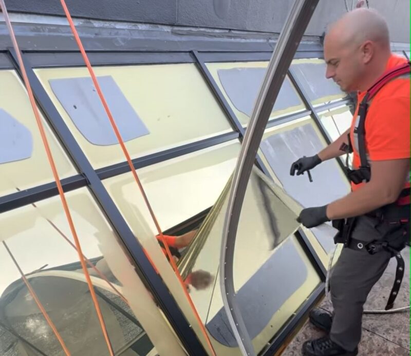 Installer in a safety harness applying window film to sloped skylight glazing on a commercial building